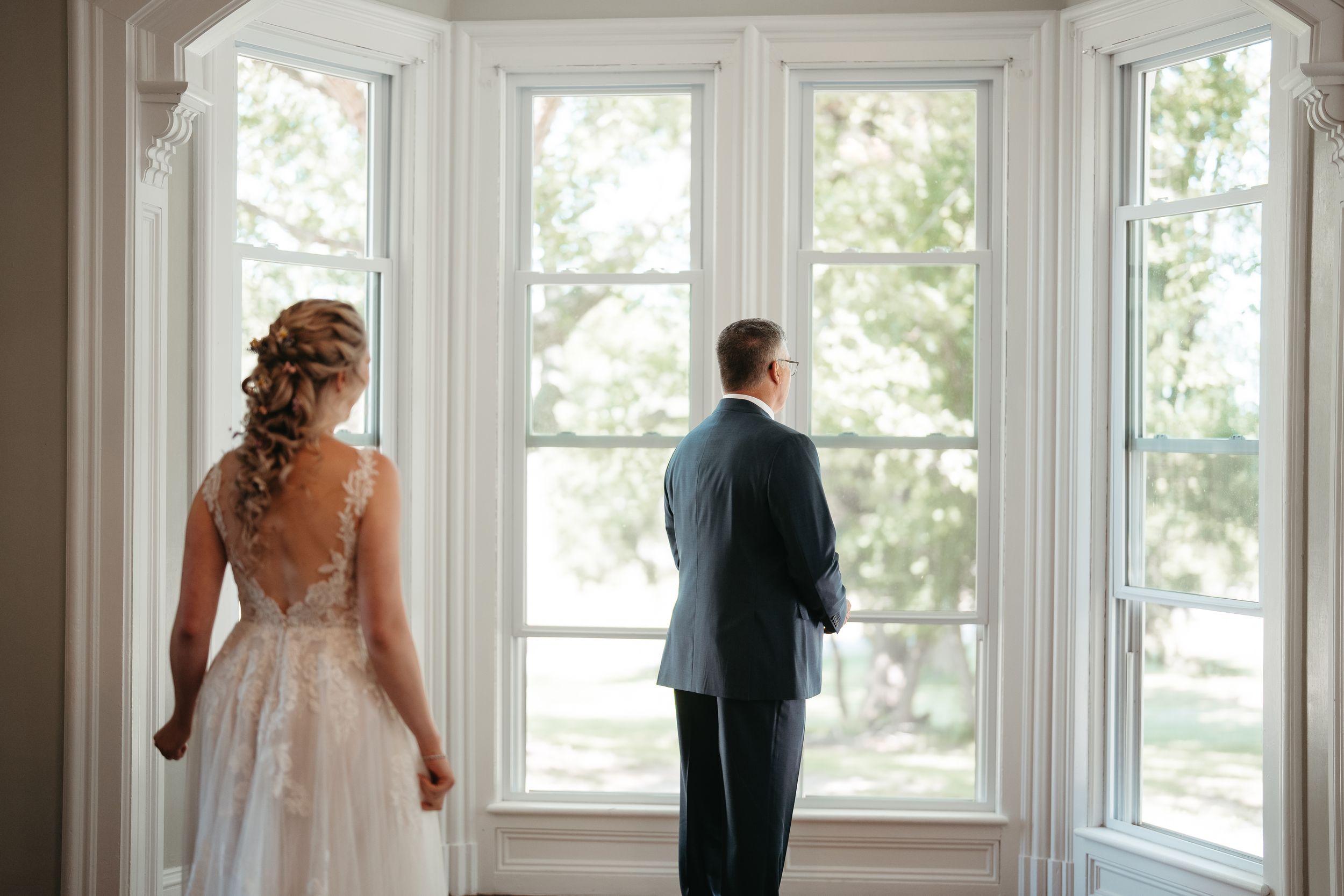 Bride and groom share a first look moment standing before large white windows with garden views on their wedding day.
