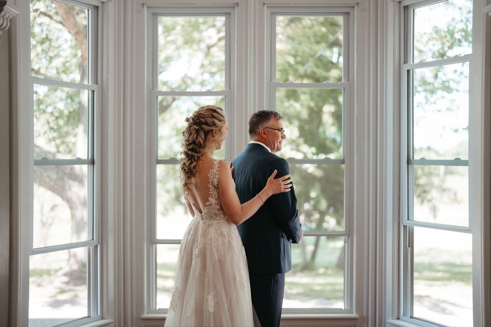 Bride and father share first look moment by large windows in elegant lace wedding gown.