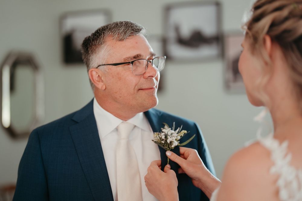 Bride pins boutonniere on older man in navy suit and glasses at wedding celebration.