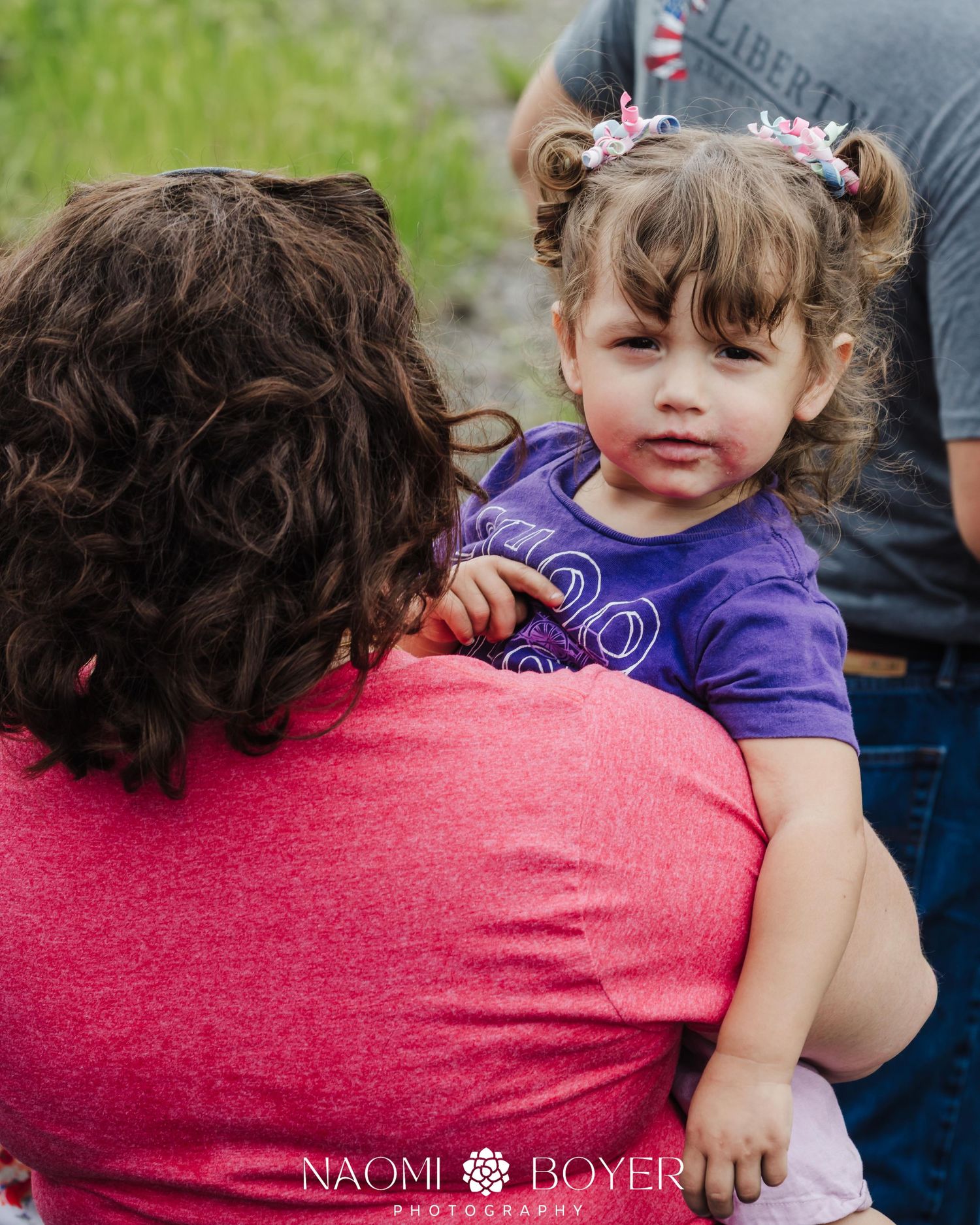 A toddler girl with pigtails in a purple shirt being held by a woman in a pink top outdoors.