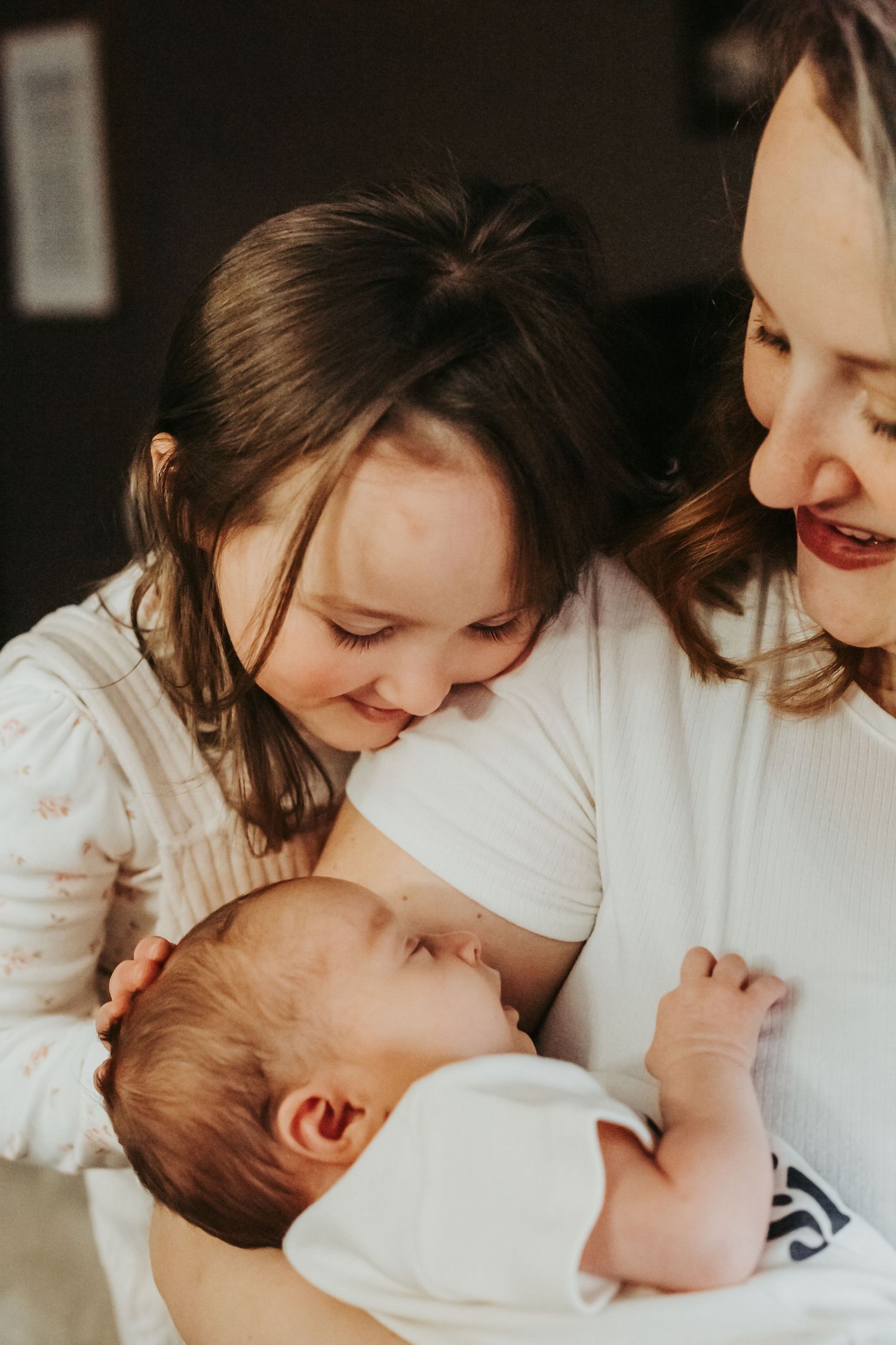 A mother and young girl smile lovingly at a newborn baby, sharing a tender family moment together.