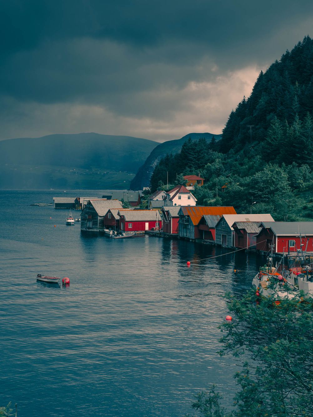 Red boathouses line a calm Norwegian fjord under dramatic stormy skies with misty mountains in the background.