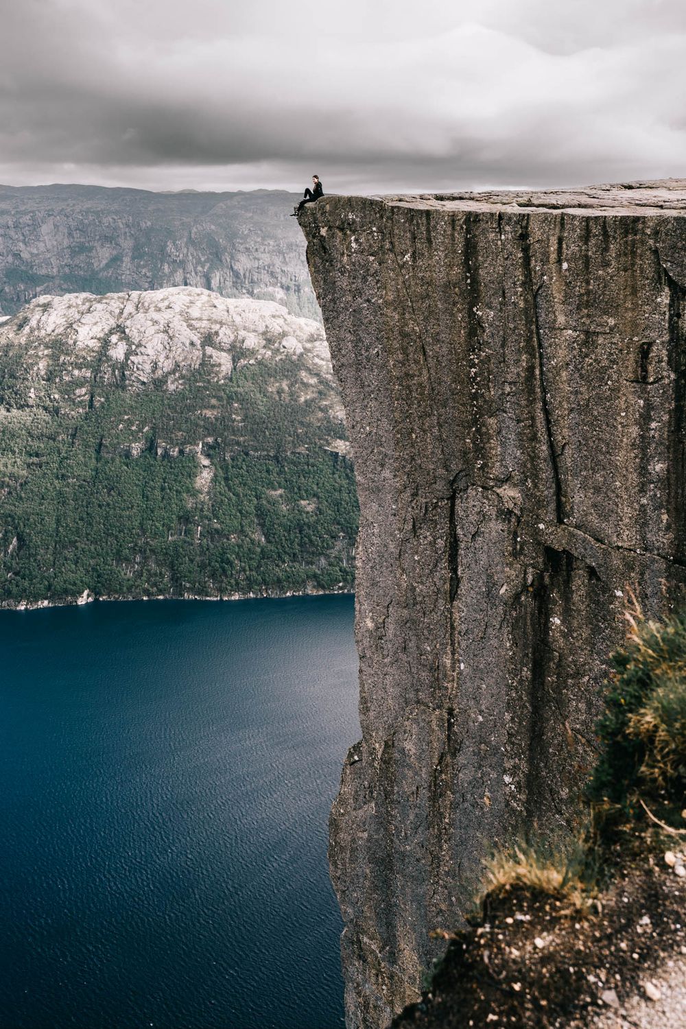 Person sitting on edge of Preikestolen cliff in Norway overlooking deep blue fjord waters below dramatic cloudy skies.