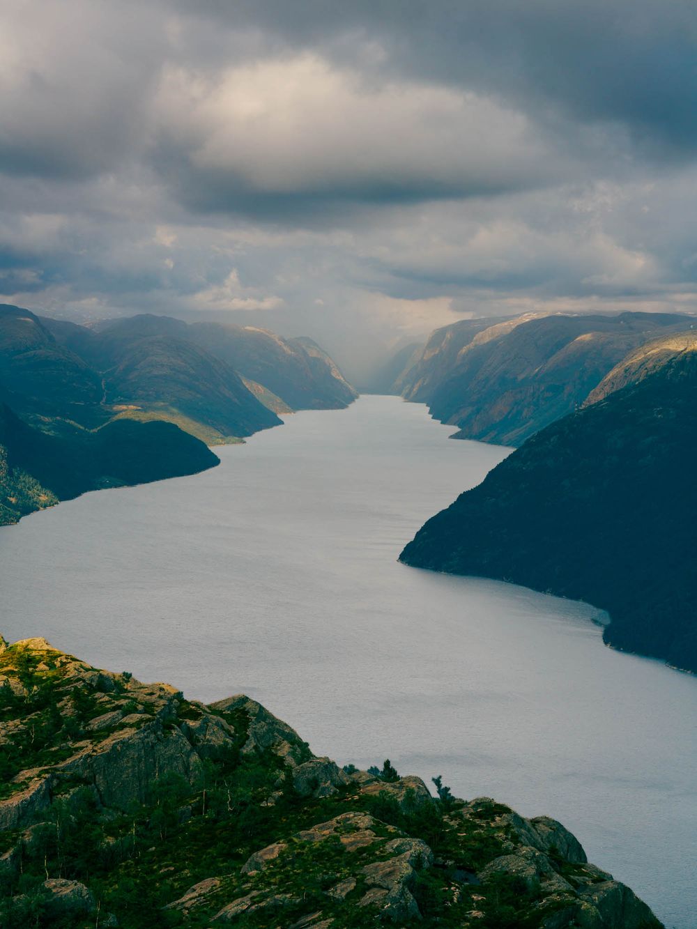 Aerial view of a serene Norwegian fjord winding between lush green mountains under dramatic cloudy skies.