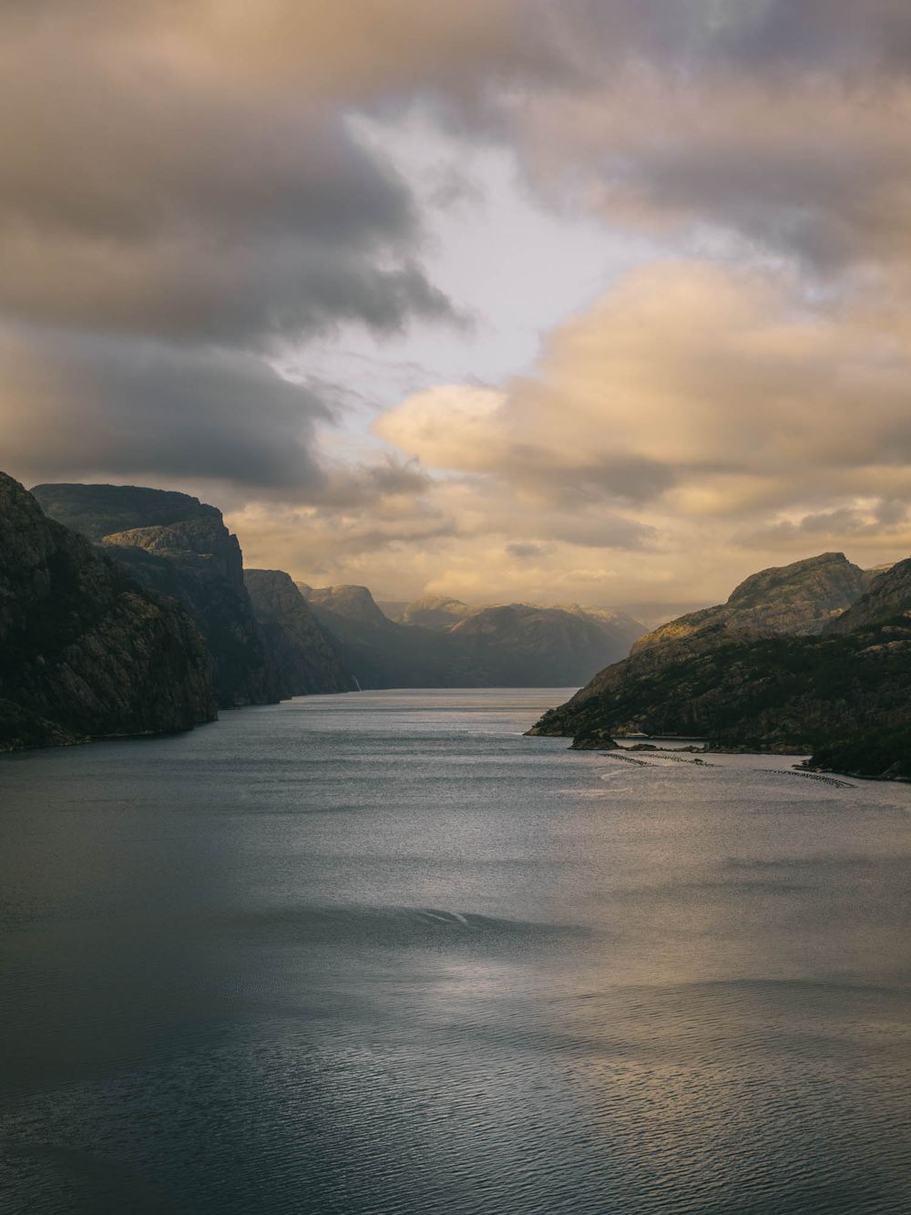 Dramatic fjord landscape with steep rocky cliffs flanking calm waters under a moody cloudy sky at golden hour.