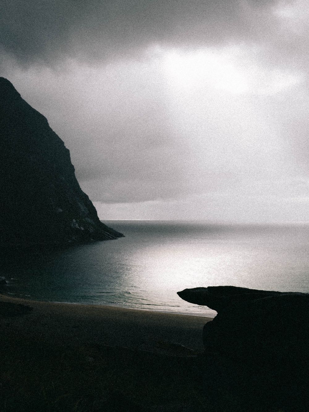 Dramatic coastal landscape with dark cliffs, calm sea, and moody overcast sky with rays of light breaking through clouds.