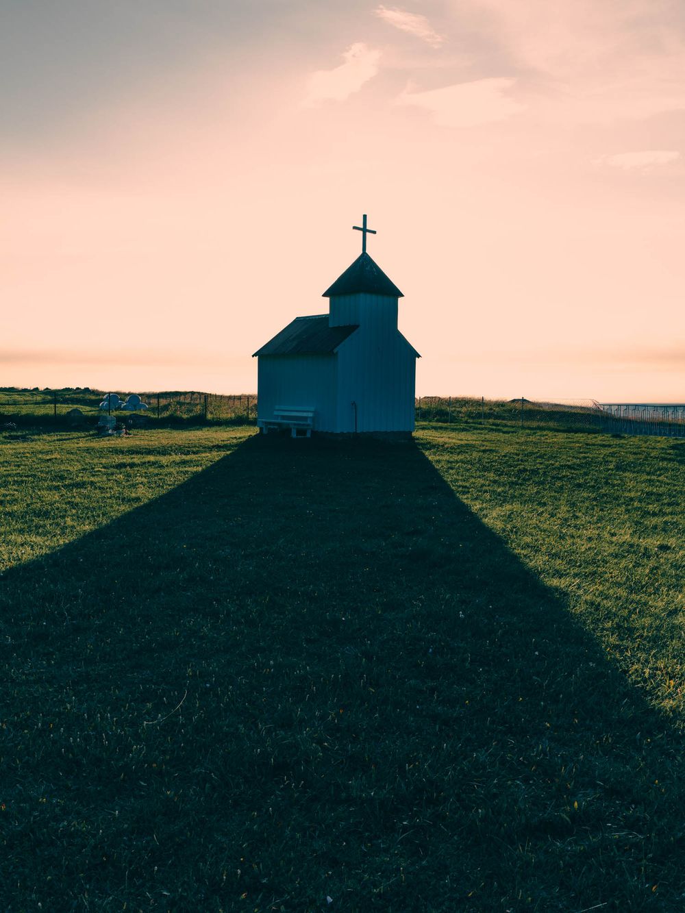 Small white chapel with a cross on a grassy hill casting a long shadow at sunset.