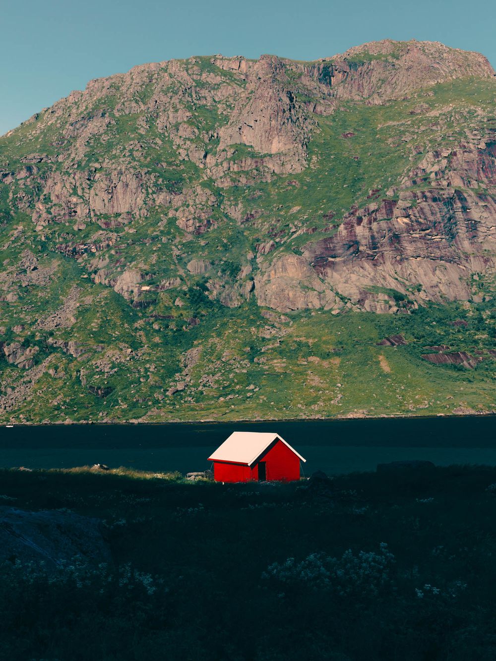A small red cabin with white roof sits beside a fjord with a dramatic green and rocky mountain backdrop in Norway.