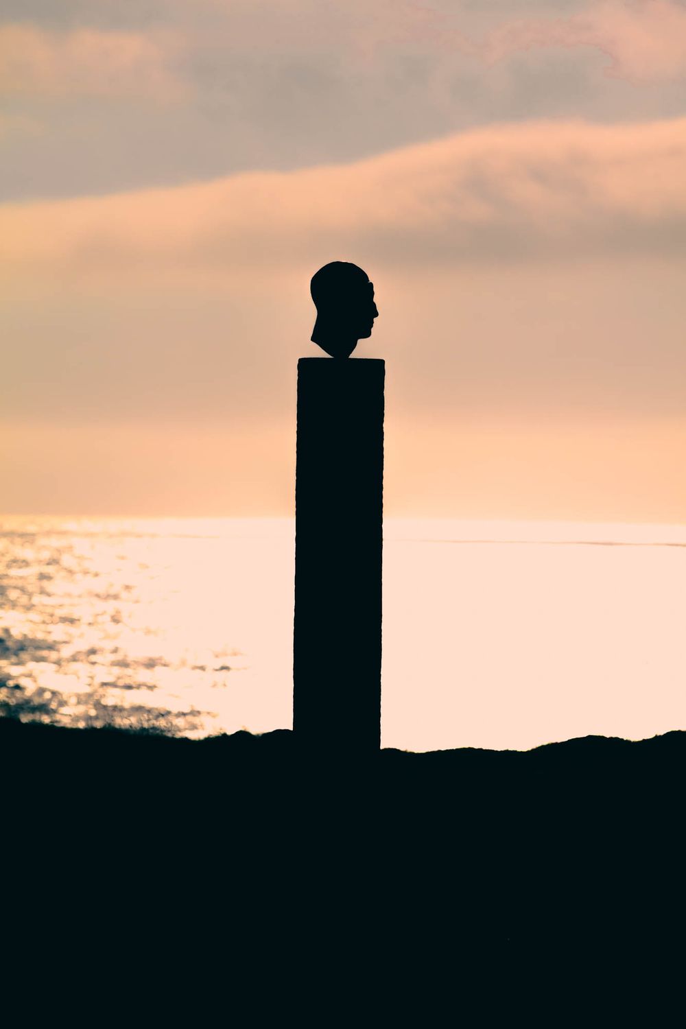 Silhouette of a bust sculpture on a tall pillar against a golden ocean sunset sky.