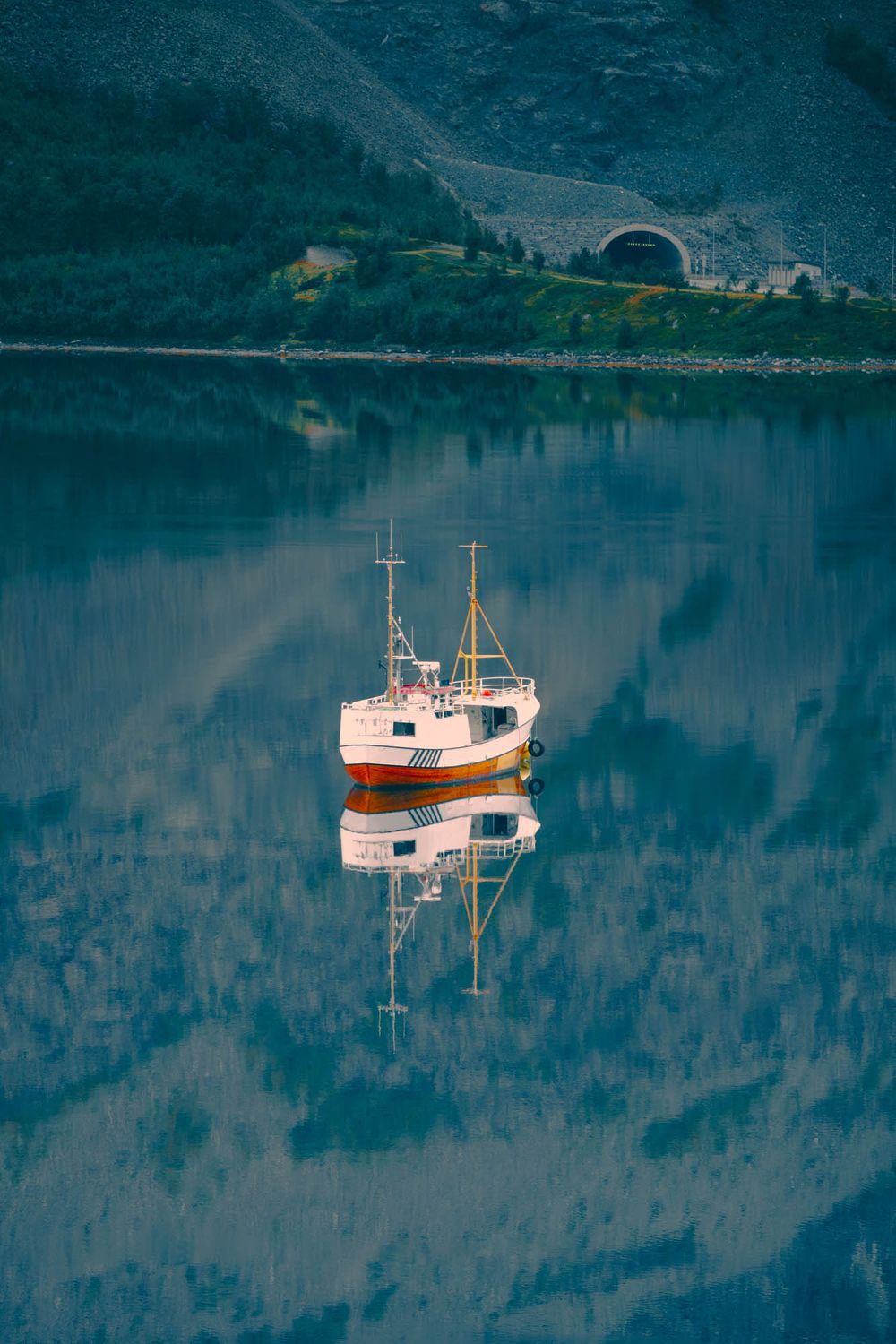 A white fishing boat reflects perfectly on the calm blue waters of a Norwegian fjord with mountains behind.