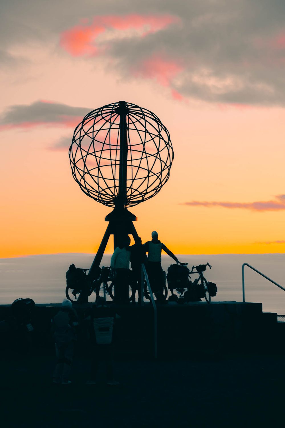 Silhouette of cyclists beside iconic North Cape globe monument against vibrant orange and pink sunset sky.