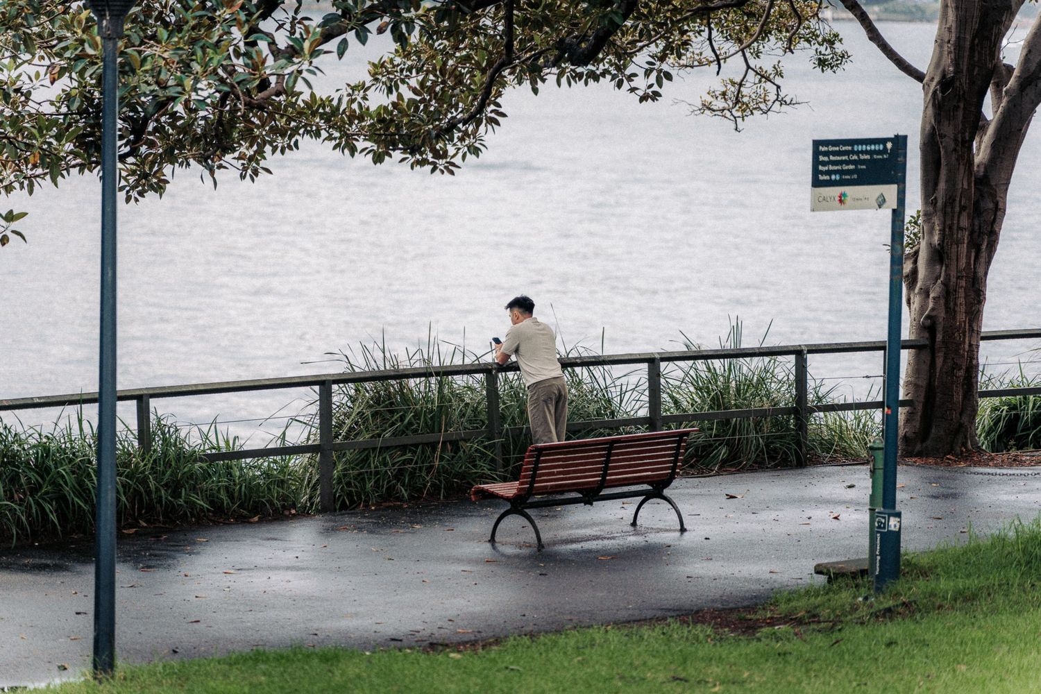 A person stands alone by a waterfront fence near an empty park bench on a overcast day.