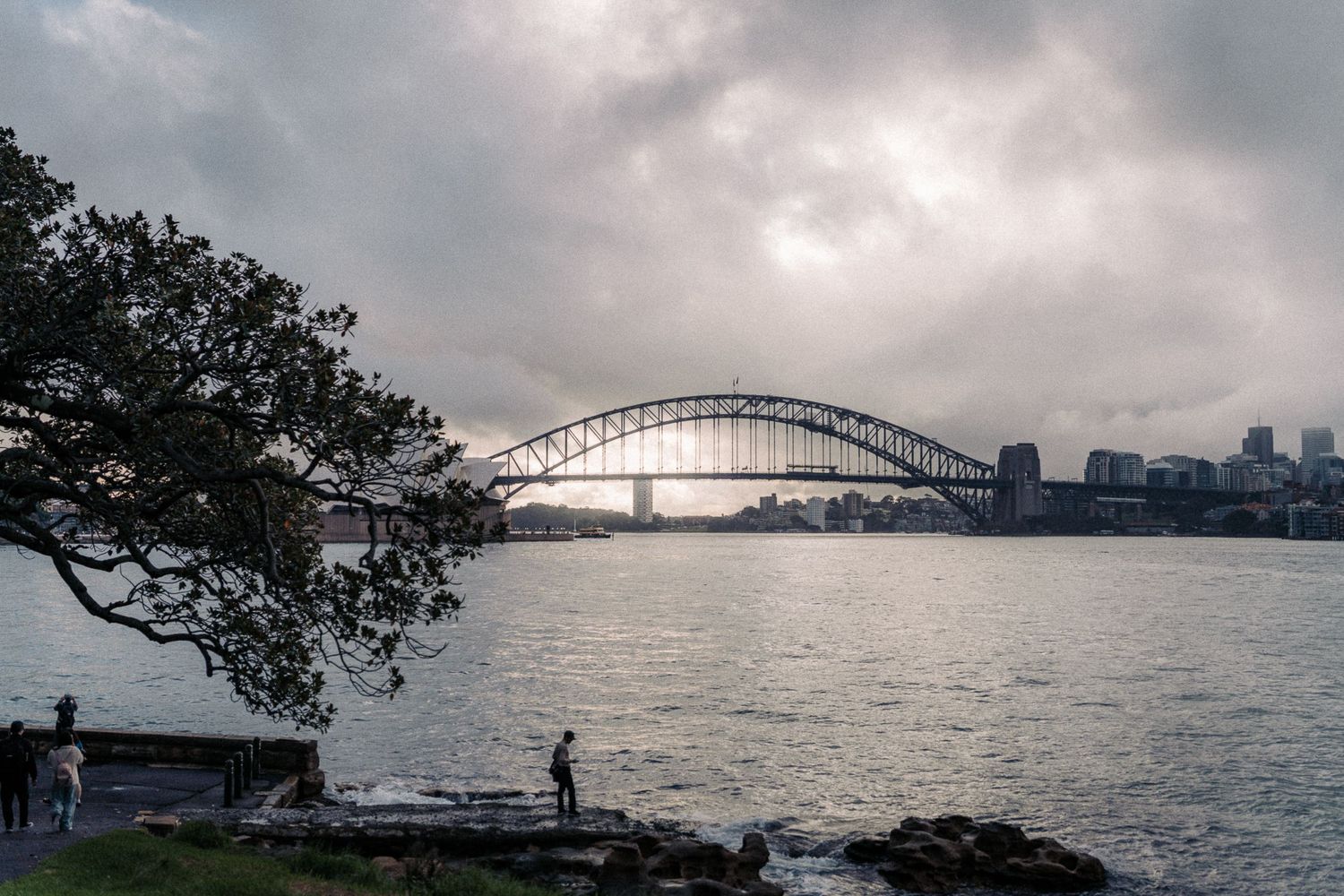 Sydney Harbour Bridge viewed from rocky shoreline under dramatic cloudy sky with city skyline in background.