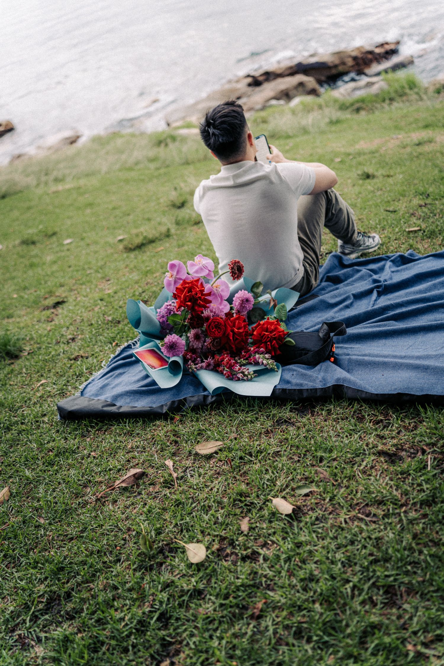 Man sitting on blue blanket by the water with a bouquet of red and pink flowers at a scenic outdoor location.