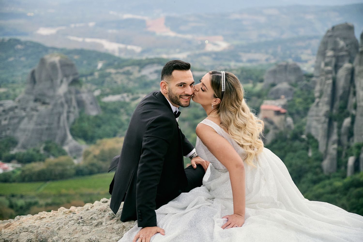 Groom and bride sitting on a cliff with the unique Meteora rock formations in the background.