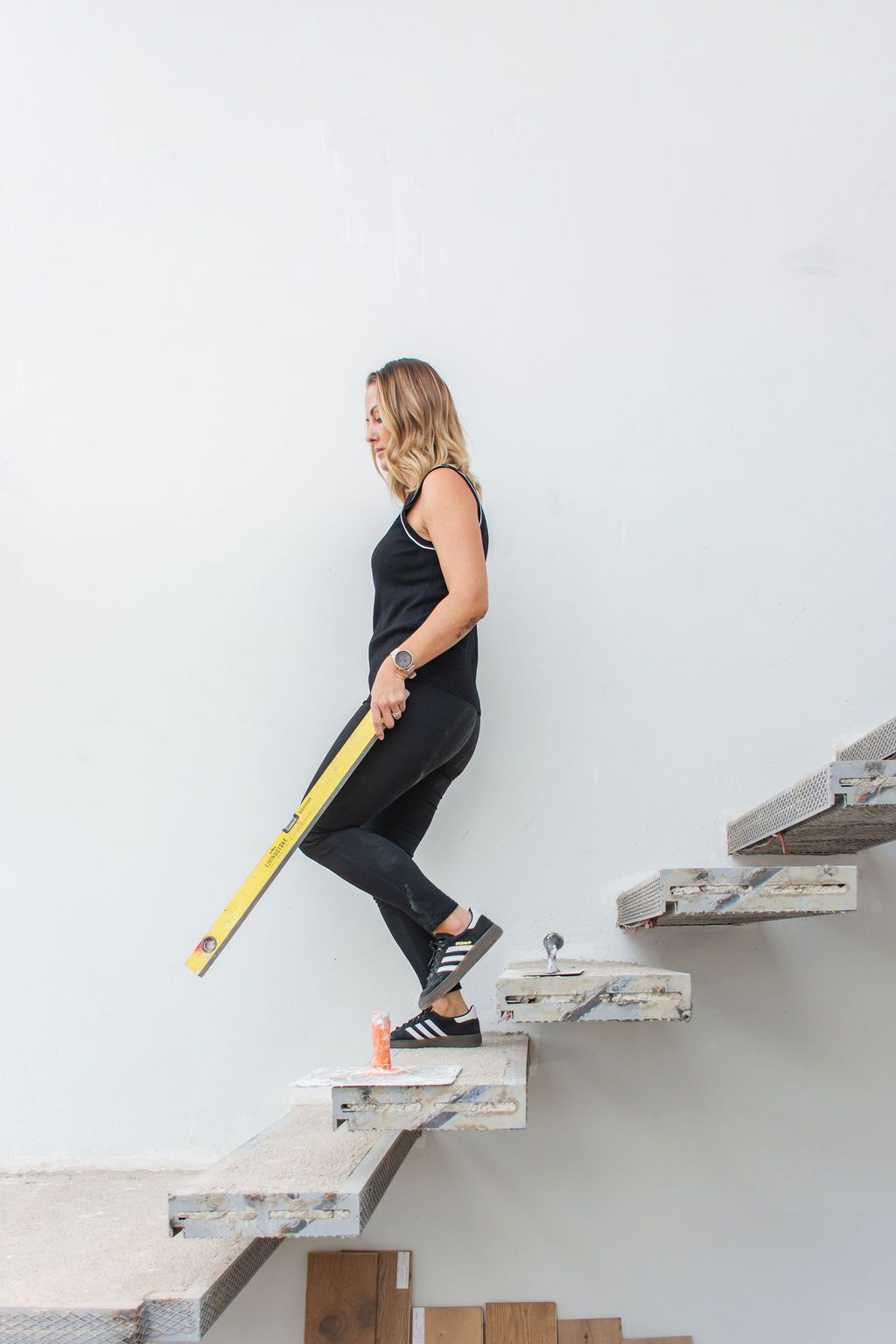 Lara, from The Goodspace Design Studio, in black athletic wear carrying a yellow level tool while walking down concrete stairs.