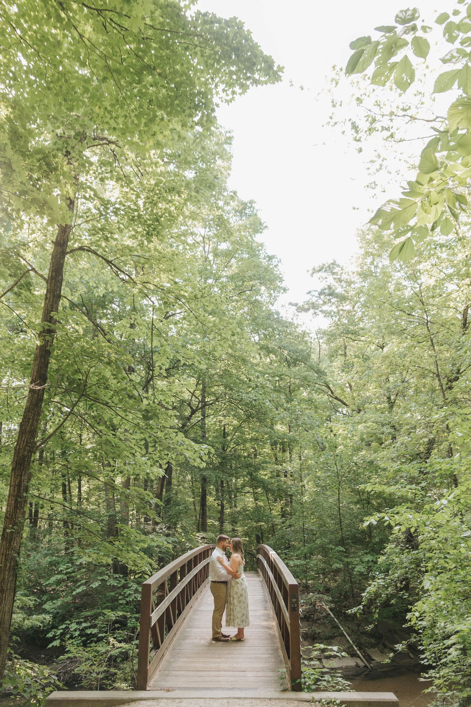 Couple embracing on a wooden bridge surrounded by lush green forest trees in a romantic outdoor setting.