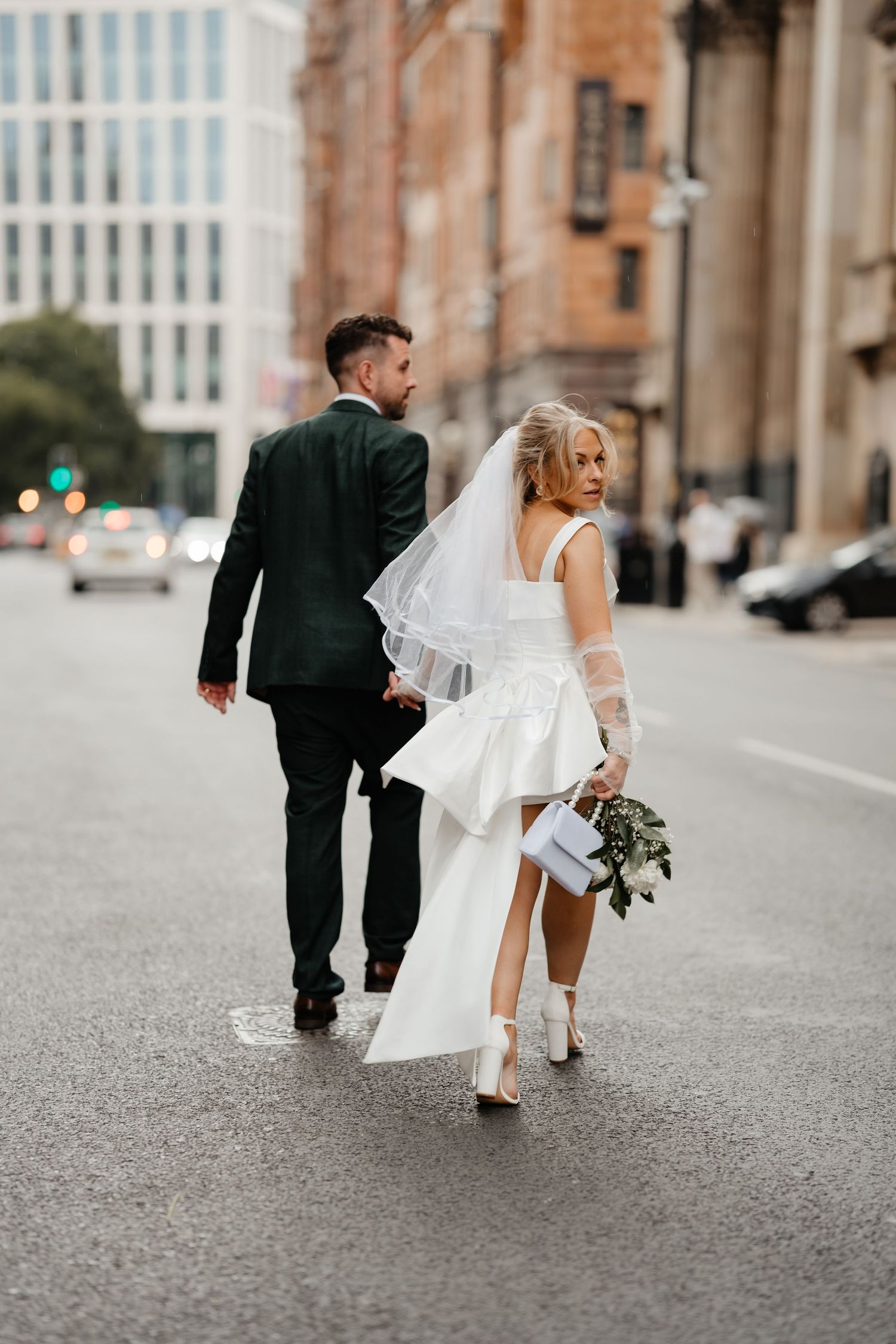 Bride and groom walking hand in hand down a city street, bride wearing short white dress with veil and bouquet.