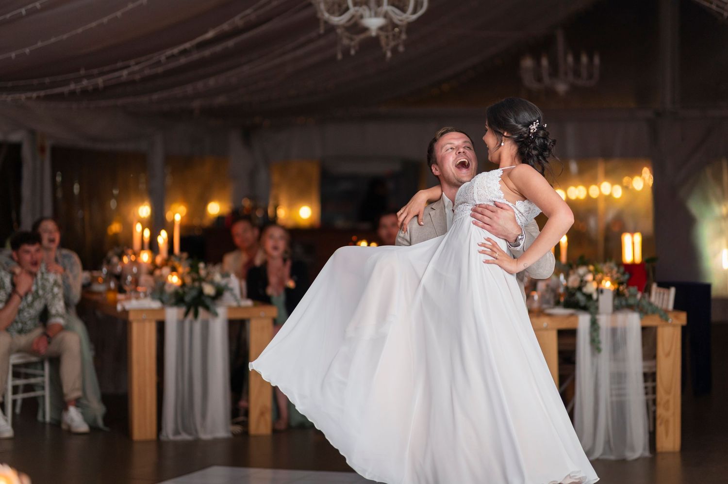 Groom joyfully lifts bride in white gown during first dance at elegant candlelit wedding reception.