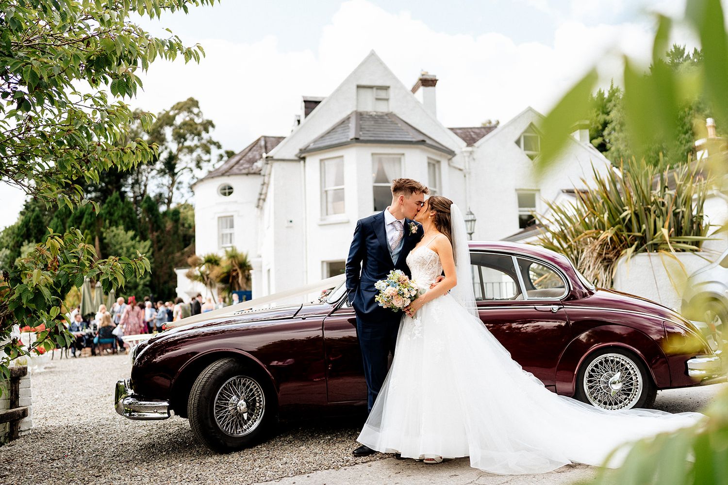 Elegant couple embraces by vintage burgundy car outside white mansion wedding venue on sunny day.