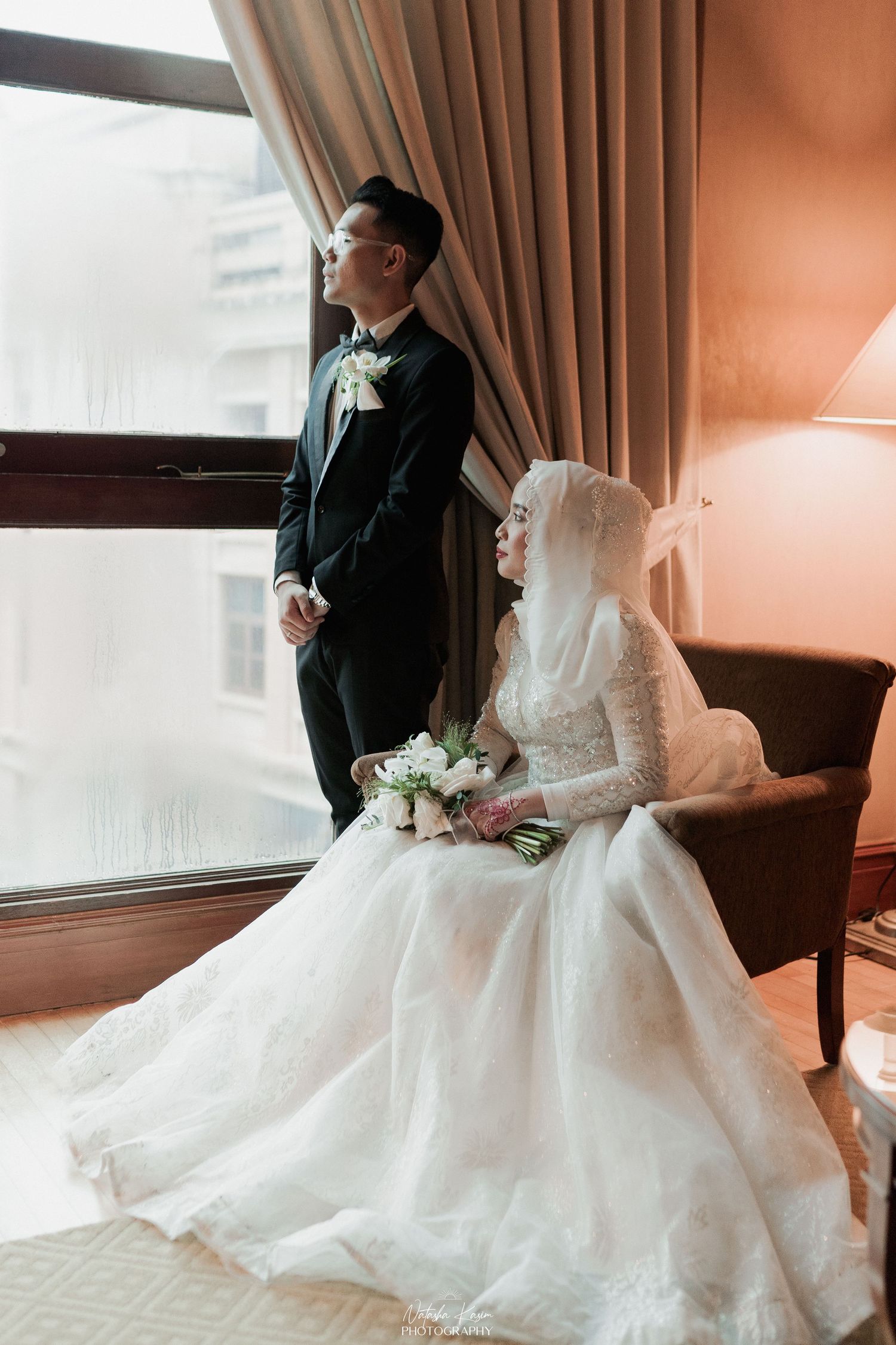 A Muslim couple in white wedding attire share a tender moment, the bride smiling in hijab as the groom leans close.