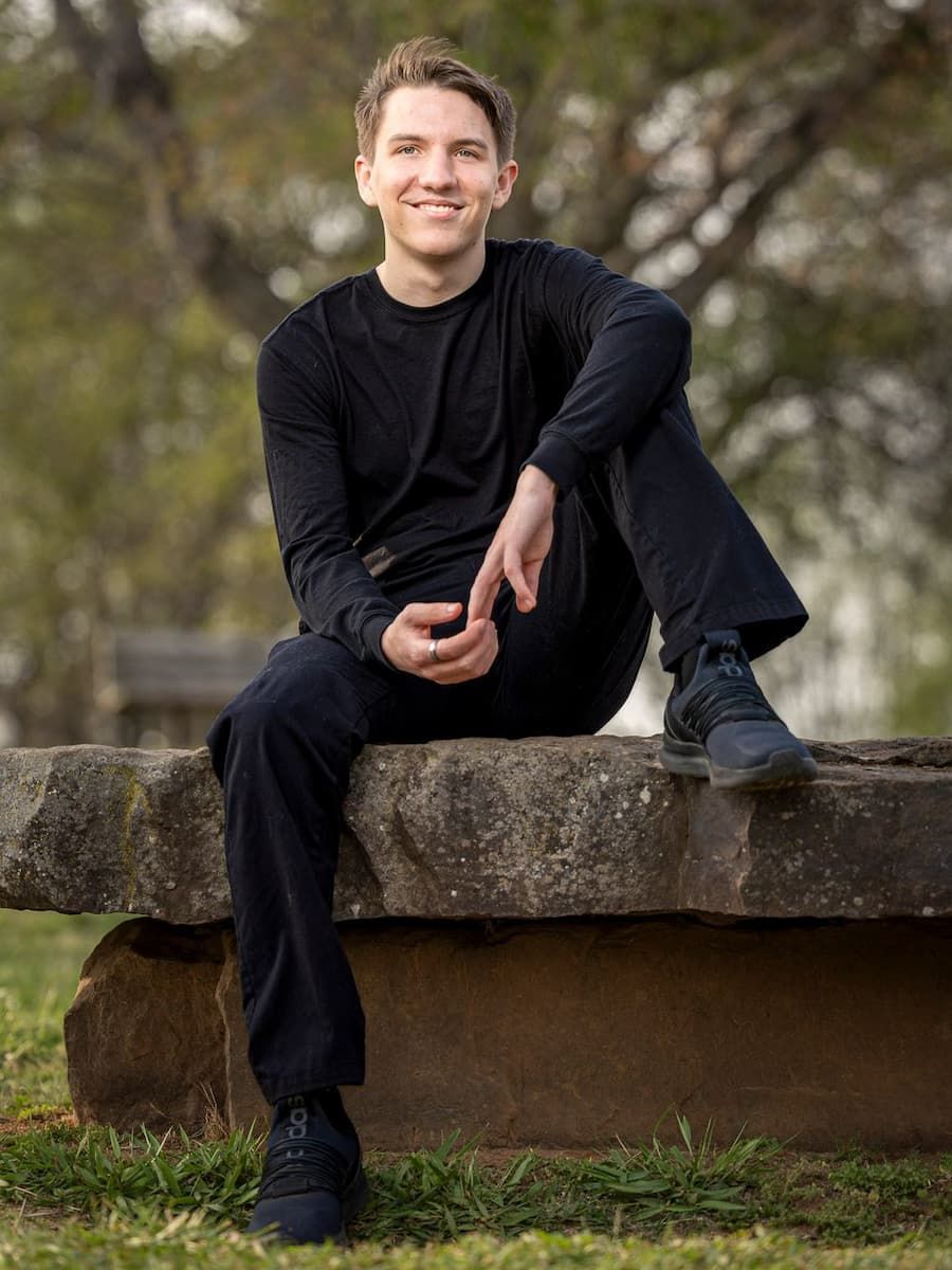 Young man in all-black outfit sitting on a stone bench outdoors, smiling confidently in a park setting.