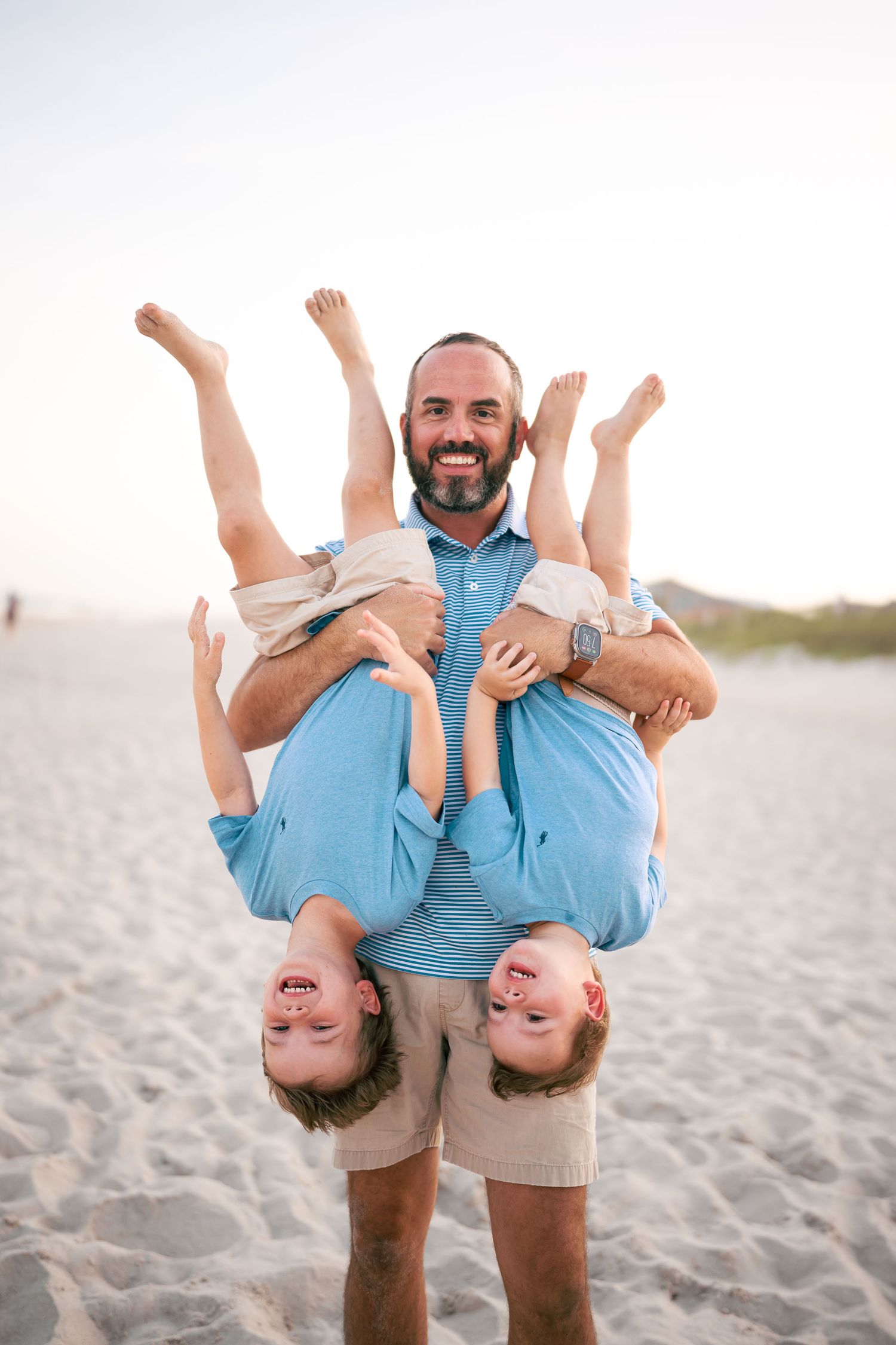 Smiling bearded father holds two laughing boys upside down on a sunny beach.