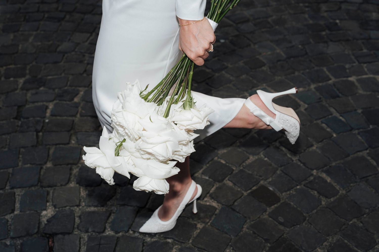 Bride in white dress holding white tulip bouquet, wearing elegant white heels on cobblestone street.