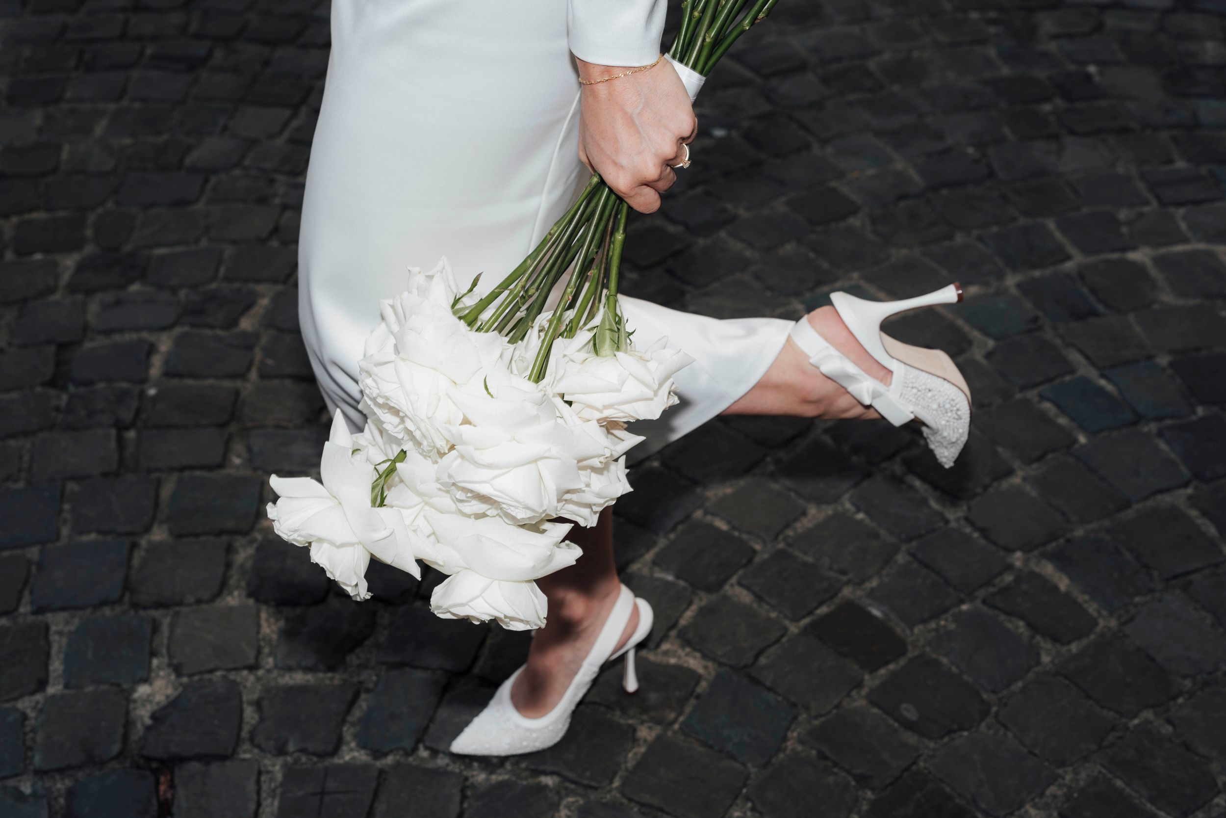 Bride in white dress holding white tulip bouquet, wearing elegant white heels on cobblestone street.