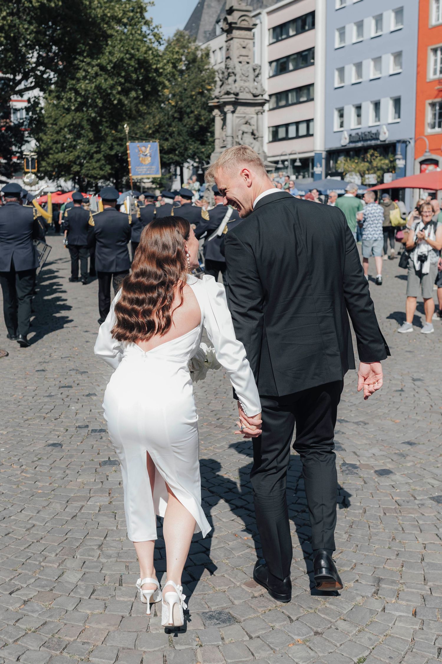 A bride in a white dress and groom in a black suit walk hand in hand through a sunny European town square.