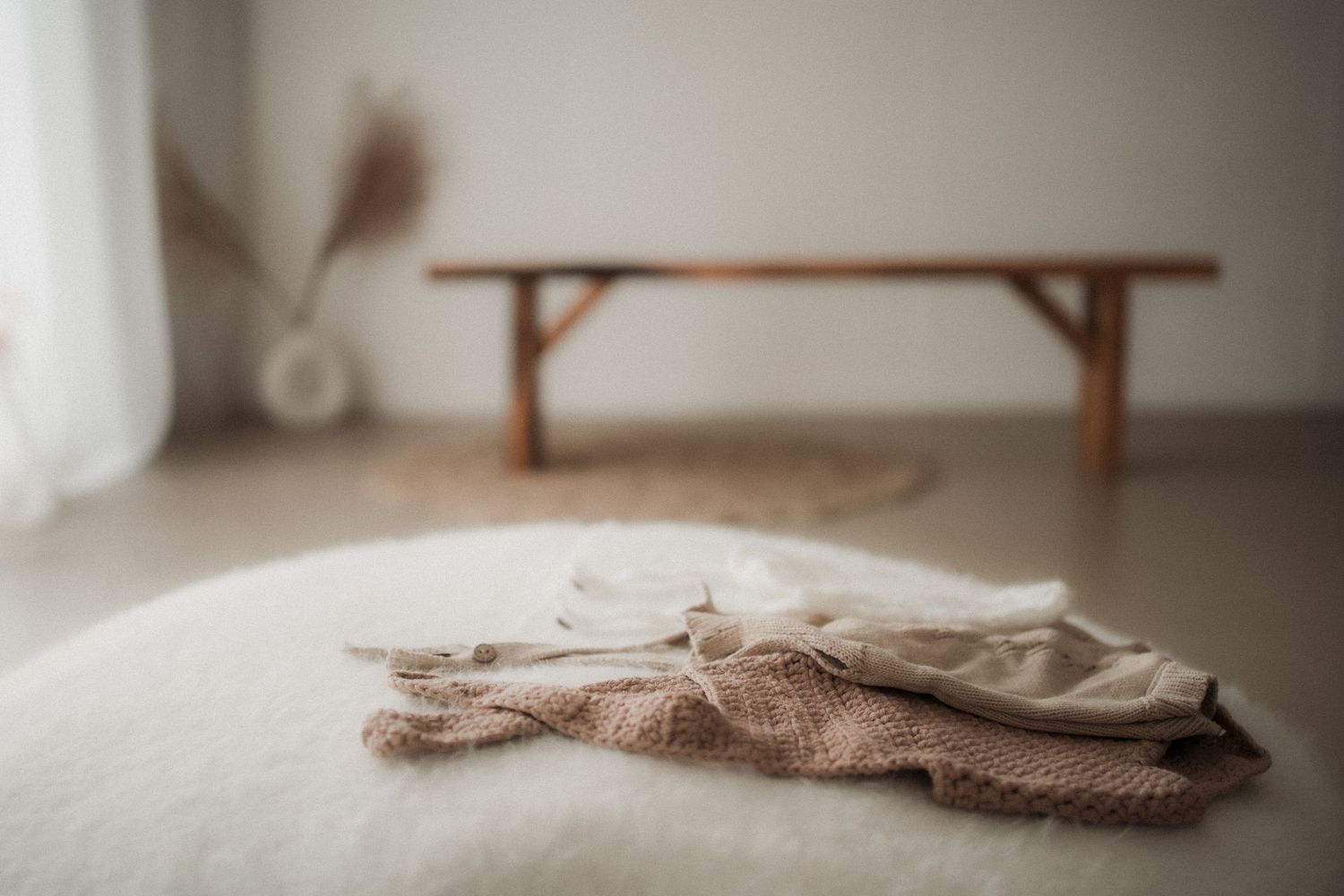 Soft knit baby clothes folded on a fluffy white rug in a minimalist room with a wooden bench in Yarra Valley Newborn Studio