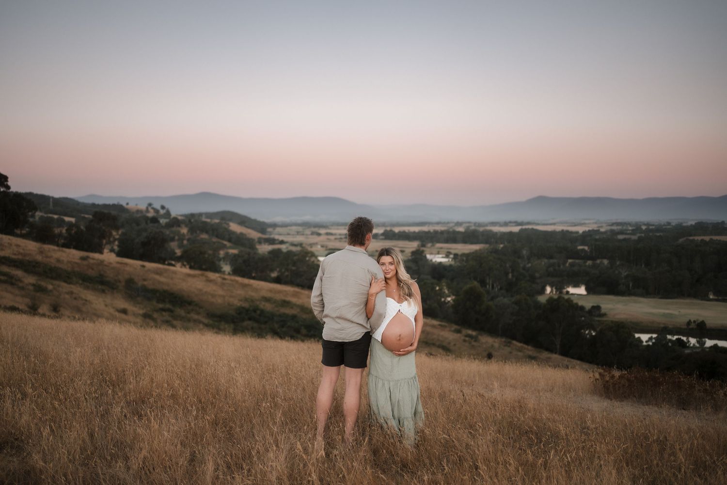 Couple standing on golden hillside at sunset with panoramic valley and mountain views during maternity photoshoot in Wonga Park, Victoria