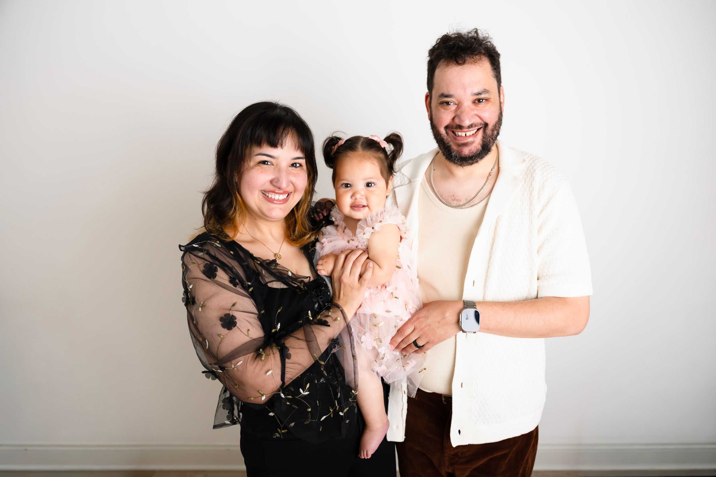A happy young family with a mother, father, and toddler daughter in pink dress posing together against a white background.