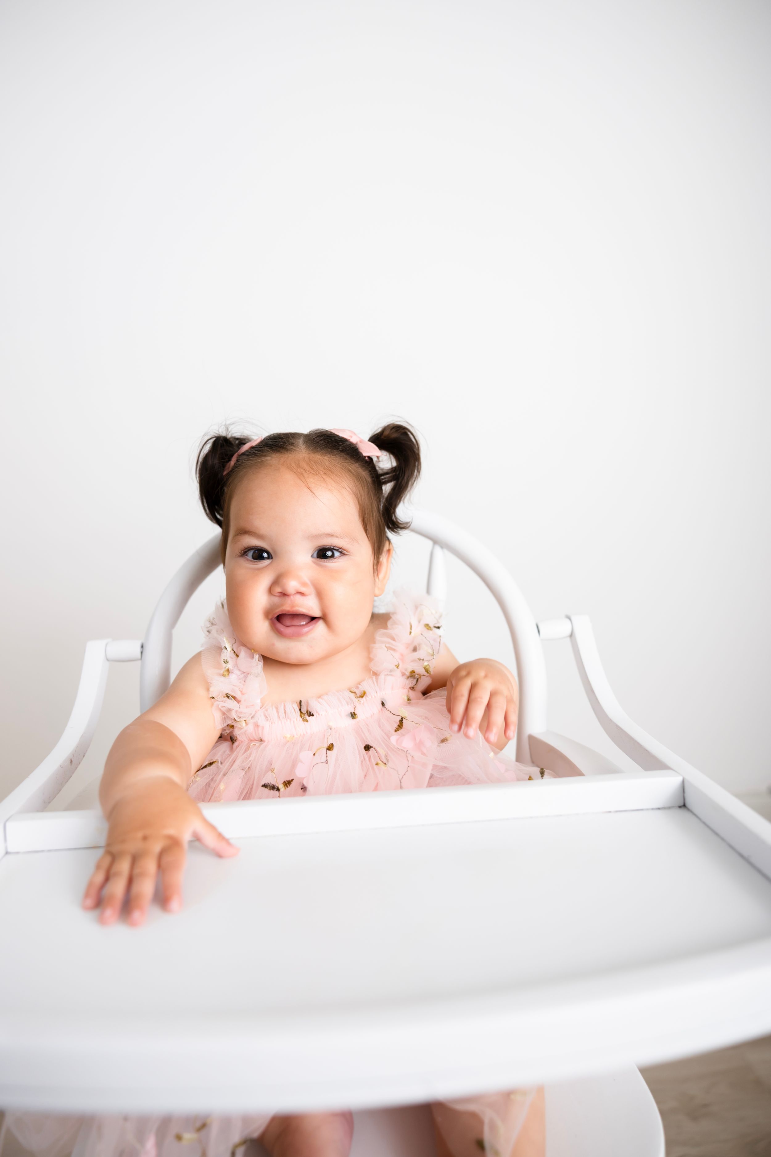 Smiling baby girl in pink dress with pigtails sitting in white high chair against white background. NJ Baby Photographer in Sussex County