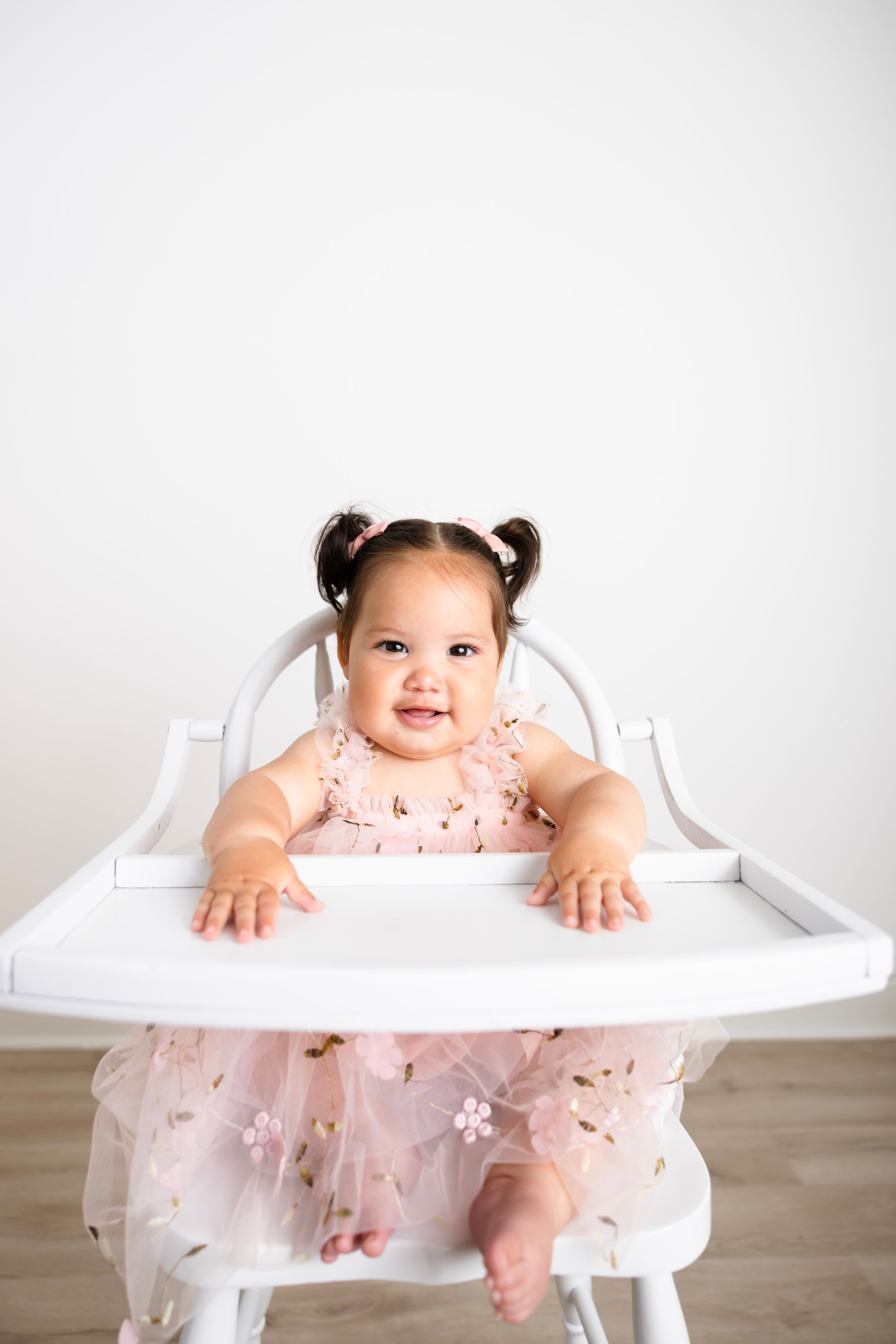 Smiling baby girl in pink floral dress sitting in white high chair against white wall.
