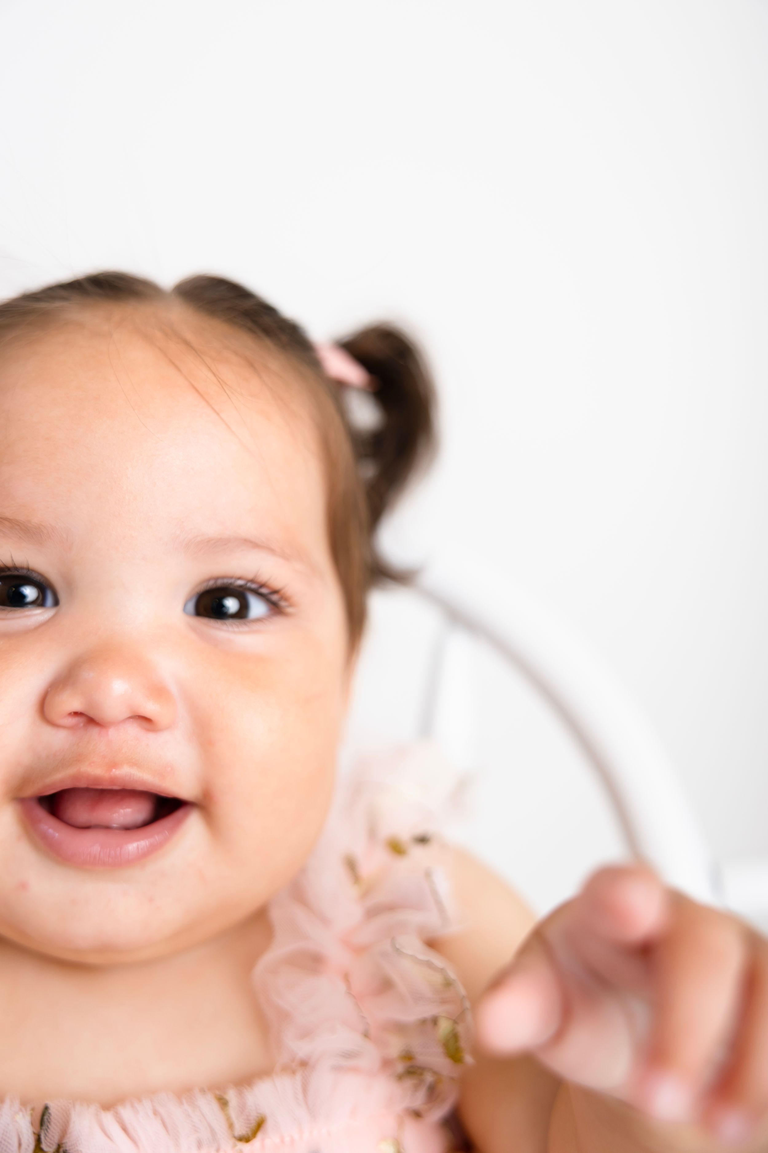 Adorable smiling baby girl with pigtails in a pink ruffle dress, showing tiny teeth in a cheerful expression. NJ Baby Photographer