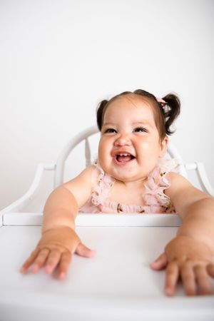 Laughing baby girl with pigtails in pink ruffled outfit sitting in white high chair reaching forward. Milestone Session in Studio Photograph
