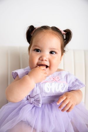 Cute baby girl in purple tutu dress with “one” text eating finger food in white chair.