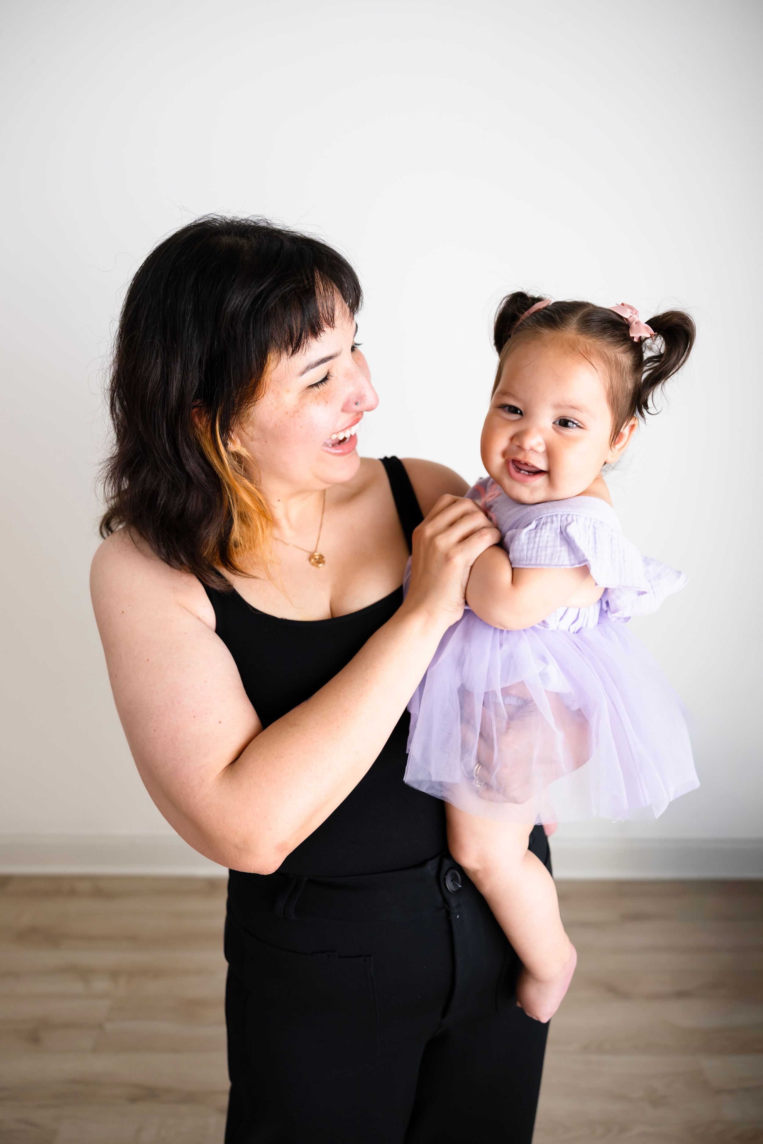 A smiling mother in a black outfit holds her happy baby girl in a purple tutu dress in a bright studio.