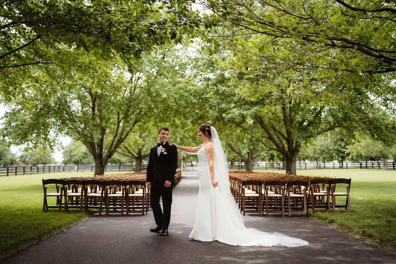 Bride and groom stand together on a tree-lined path with wooden ceremony chairs set up outdoors on a lush green lawn.