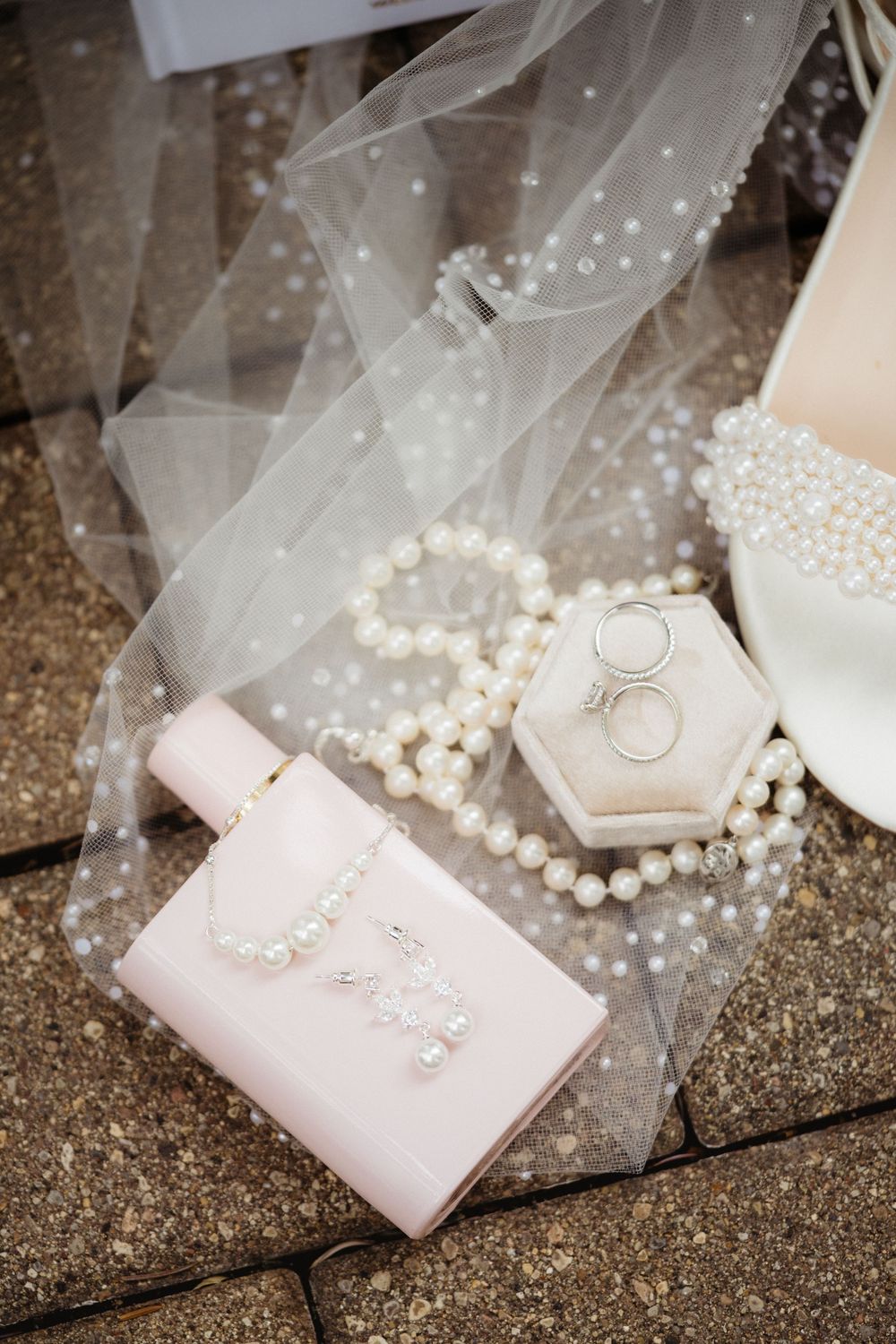 Bridal accessories flat lay with pearl jewelry, wedding rings in velvet box, pink clutch, veil, and pearl-embellished heels.