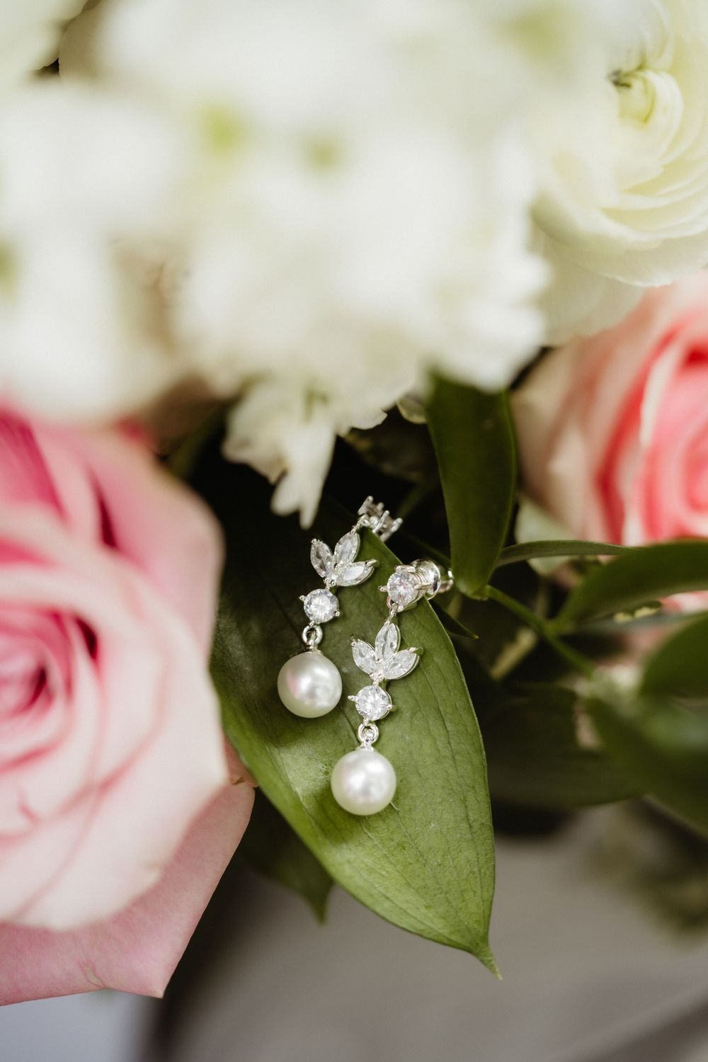 Pearl and crystal drop earrings resting on green leaves surrounded by pink and white roses in a bridal bouquet.
