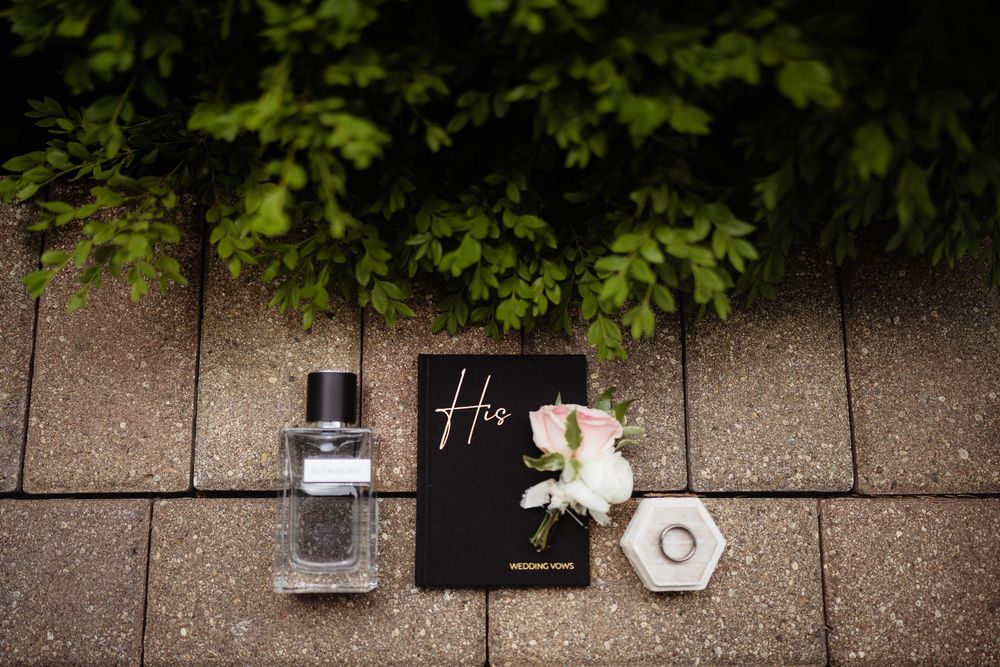 Groom wedding flat lay with cologne, black vow book, white rose boutonniere, and ring box on stone pavers.