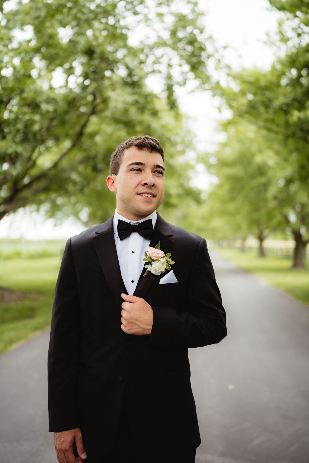 Groom in black tuxedo with bow tie and floral boutonniere standing on a tree-lined path at an outdoor wedding.