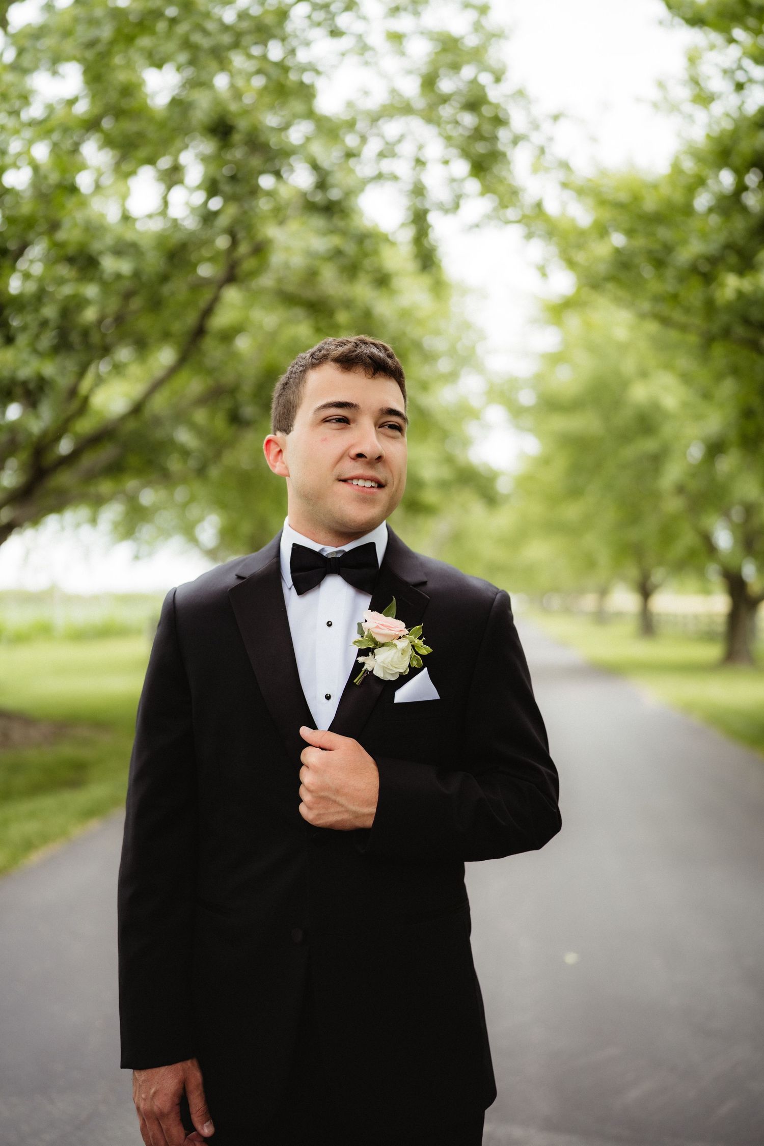 Groom in black tuxedo with bow tie and floral boutonniere standing on a tree-lined path at an outdoor wedding.