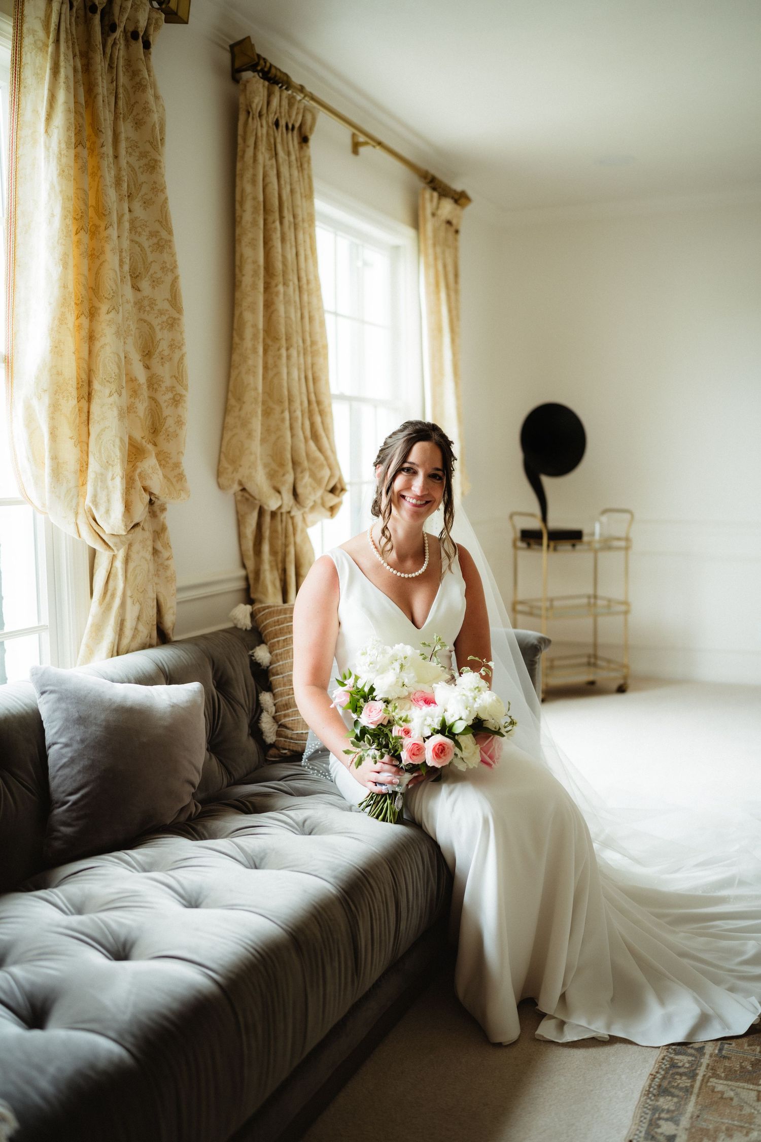 Bride in white gown sits on gray tufted sofa holding pink and white bouquet, smiling near sunlit window with gold curtains.
