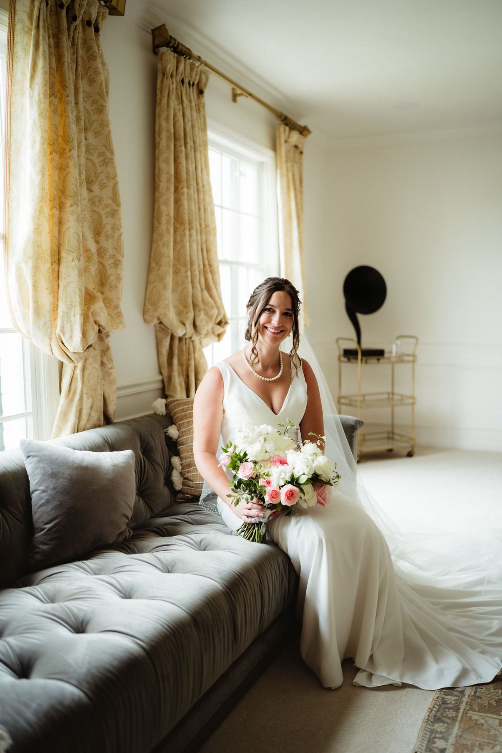 Bride in white gown sits on gray tufted sofa holding pink and white bouquet, smiling near sunlit window with gold curtains.