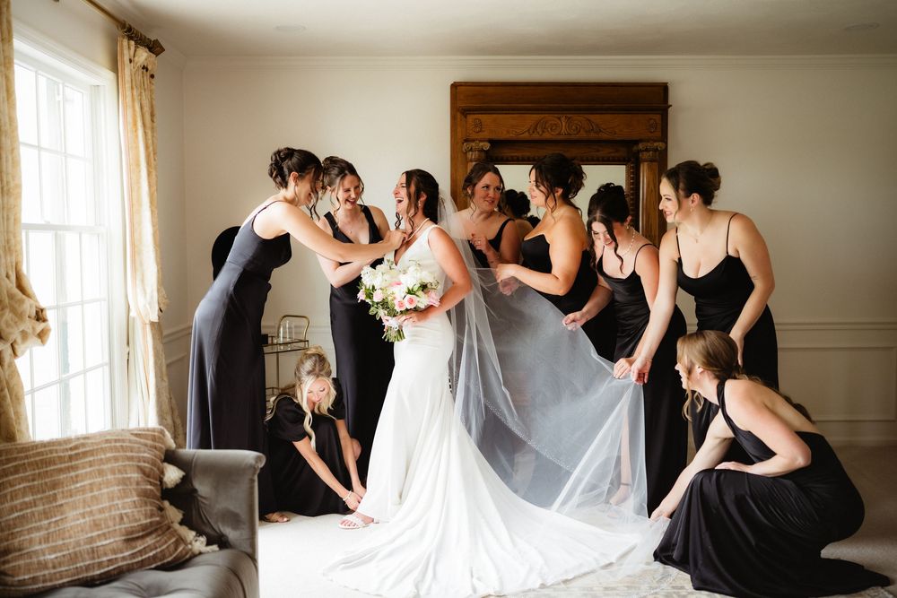 Bridesmaids in black dresses help bride with white gown and veil in bright room before wedding ceremony.