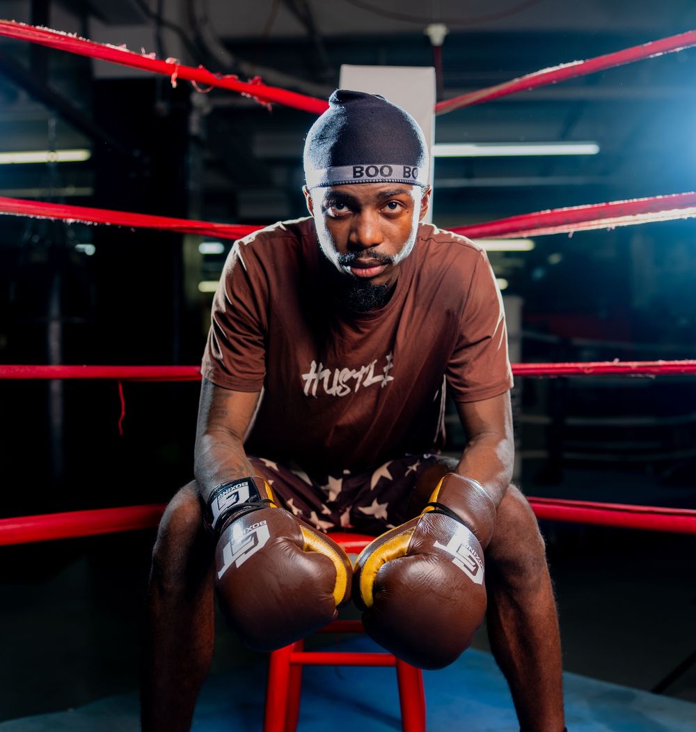 Boxer in brown Hustle shirt and beanie sits on stool in boxing ring wearing gold gloves.