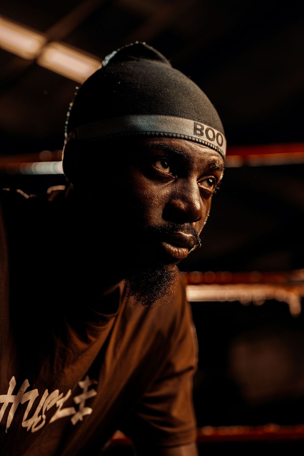 A focused male boxer wearing a black beanie and Hustle shirt rests in a boxing ring under dramatic lighting.